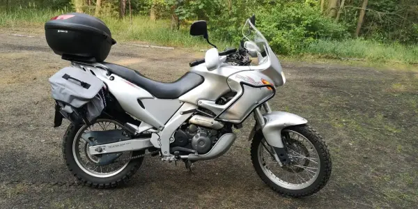Srebrny motocykl Aprilia Pegaso 650 z kufrem i sakwami, gotowy na przygodę w lesie.