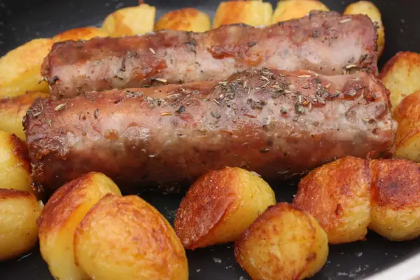 Deux andouillettes artisanales dorées, parsemées d'herbes, mijotent avec des pommes de terre rôties dans une poêle.
