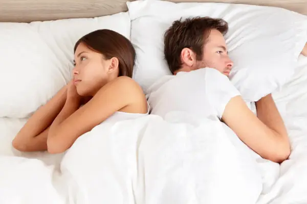 Un couple allongé dos à dos dans un lit blanc, chacun perdu dans ses pensées, un soupir amoureux flotte dans l'air.