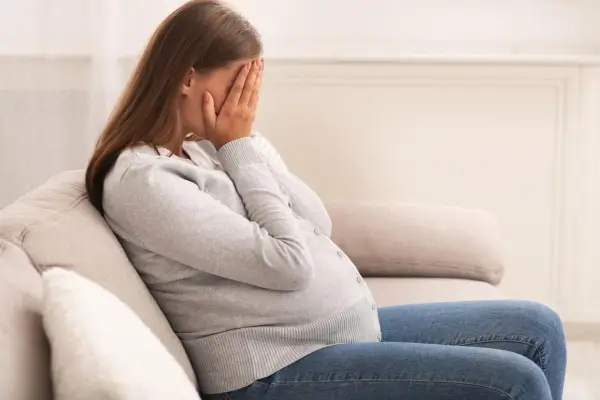 Kobieta w ciąży siedzi na kanapie, zakrywając twarz dłońmi. Jej postawa wyraża głęboki smutek, być może związany z diagnozą wady letalnej.