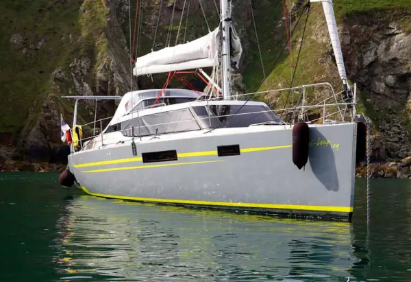 Le voilier "Bi-loup 109" gris avec des bandes jaunes, ancré près d'une falaise verdoyante.