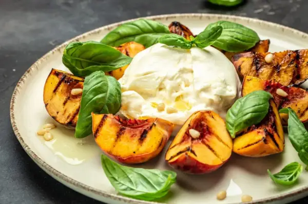 Burrata z grillowanymi brzoskwiniami, bazylią i orzeszkami piniowymi na talerzu.