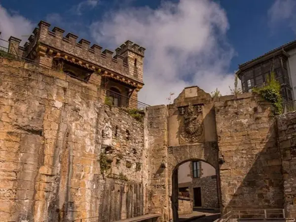 Antigua muralla y arco de entrada en Fuenterrabía, con un escudo nobiliario y un cielo azul con nubes.