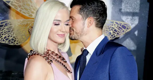 Orlando Bloom szepcze coś do ucha Katy Perry, która ma na sobie różową sukienkę i gruby, złoty naszyjnik.