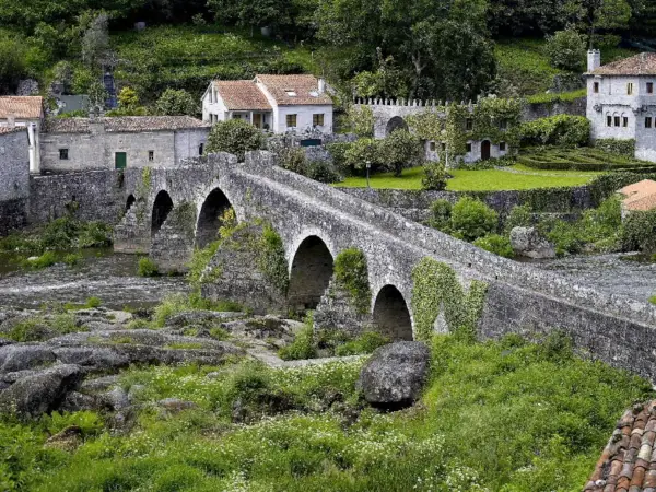 Puente de piedra cubierto de hiedra, con casas tradicionales al fondo. Un lugar idílico para visitar, así es como ponte Maceira como llegar.