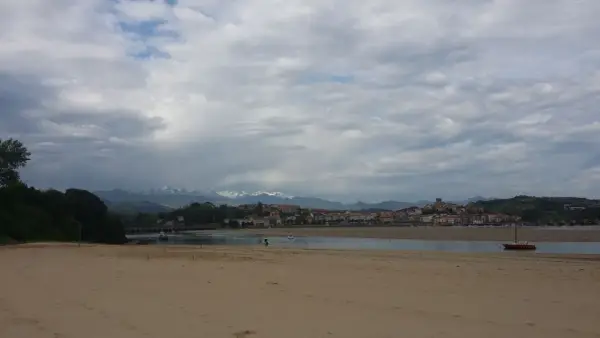 Arena dorada en la playa de la Maza, con un velero rojo en el agua y un pueblo costero al fondo, bajo un cielo nublado.