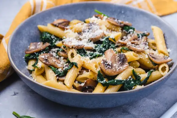 Penne z pieczarkami i szpinakiem, posypane parmezanem, podane w szarej misce.