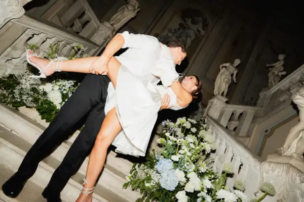 Un couple s'embrasse passionnément lors de leur mariage chez soi, entouré de fleurs blanches et bleues sur un escalier orné de statues.