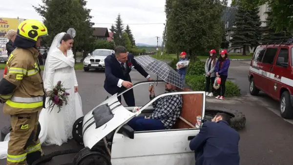 Brama weselna z udziałem strażaka, pana młodego i panny młodej. Pan młody rozmawia z druhem siedzącym w prowizorycznym samochodzie.