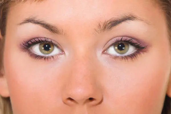 Primo piano di un viso femminile con occhi piccoli e verdi, ciglia lunghe e trucco viola sulle palpebre.