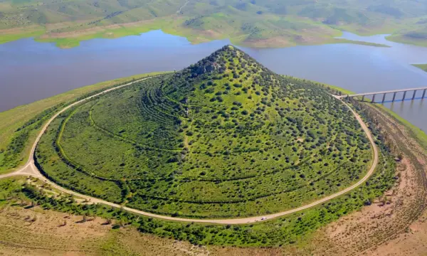 El **cerro Masatrigo**, cubierto de vegetación, se alza majestuoso junto a un embalse. Una carretera serpentea a su alrededor, conectando con un puente.