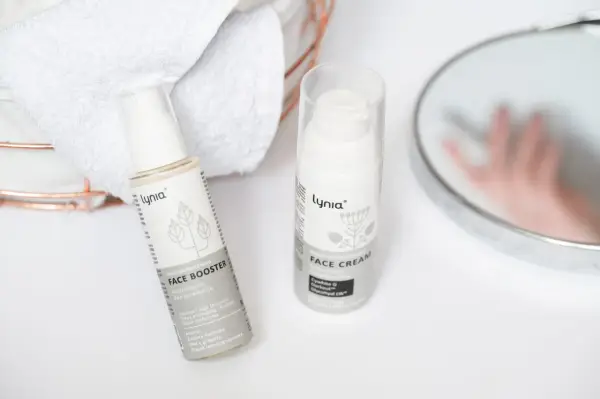 Białe opakowania kosmetyków Lynia: Face Booster i Face Cream, z widocznym składnikiem papaina. Obok ręczniki i lustro.