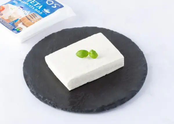 Un blocco di feta DOP su una pietra scura, guarnito con basilico. La confezione è visibile sullo sfondo.