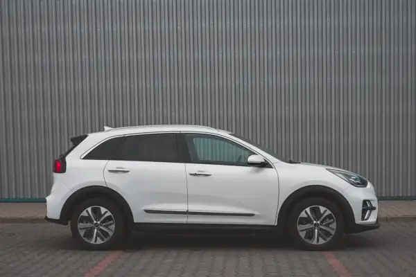 Biały Kia Niro EV stoi na tle szarej, falistej ściany. Elegancki design i nowoczesne linie podkreślają jego ekologiczny charakter.