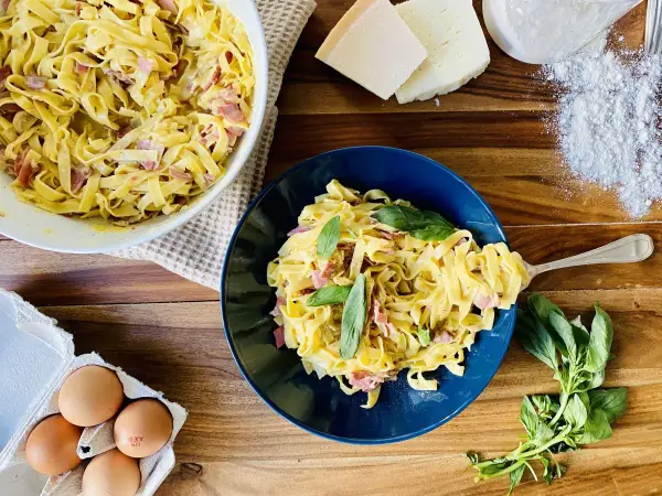 Un plat de pâtes à la carbonara crémeuses, garni de basilic frais. Des ingrédients comme des œufs, du fromage et de la farine sont visibles à proximité.