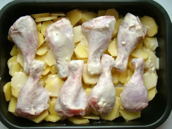 Osiem pałek kurczaka w sosie, ułożonych na plastrach ziemniaków w brytfannie. Idealne do pieczenia, by uzyskać chrupiące pałki z kurczaka w piekarniku.