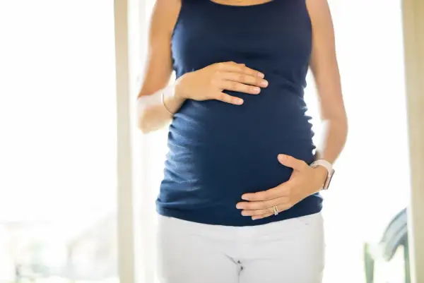 Ciężarna kobieta w granatowej bluzce i białych spodniach obejmuje dłońmi brzuch. Wody płodowe otulają rozwijające się życie.