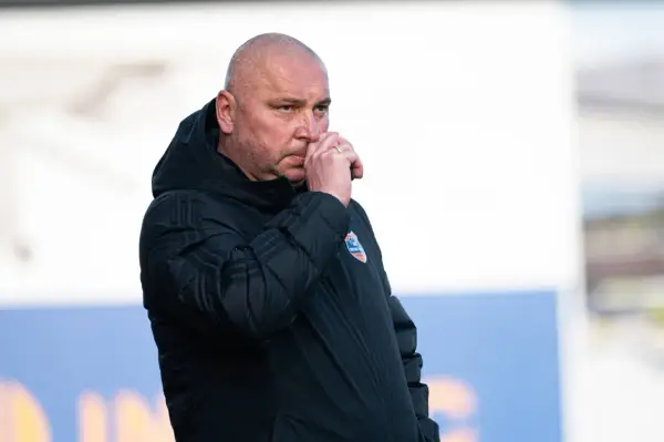 Mariusz Lewandowski, trener w czarnej kurtce, zamyślony, z ręką przy nosie.