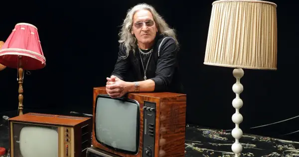 Mężczyzna z długimi siwymi włosami i okularami opiera się o stary telewizor. Wokół niego lampy i inne telewizory. Widać, że trzeba wiedzieć, kiedy ze sceny zejść.