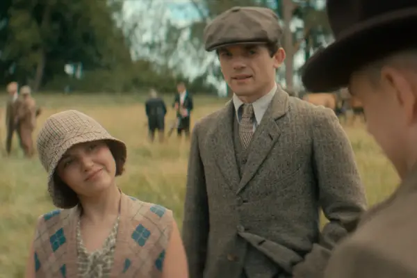 Giovane donna con cappello a scacchi e gilet a rombi, accanto a un uomo con berretto e giacca di tweed. Sullo sfondo, i sette quadranti di un campo.