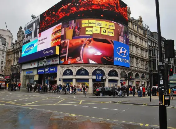 Tętniący życiem Piccadilly Circus w Londynie z neonowymi reklamami, w tym Samsung, McDonald's i Hyundai. Tłum ludzi na chodniku.