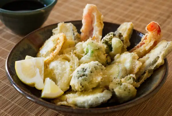 A bowl of crispy vegetable tempura, featuring broccoli, zucchini, and shrimp, served with lemon wedges and a side of dipping sauce.