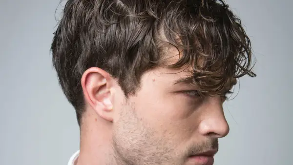 El pelo hombre, oscuro y ondulado, cae sobre su rostro. Se aprecian su oreja y parte de su barba incipiente.