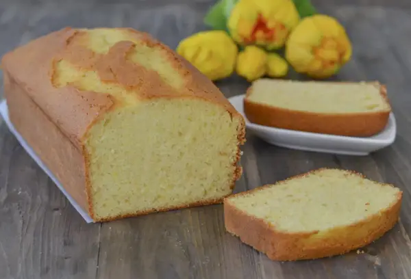 Un soffice plumcake al limone, appena sfornato, con una fetta su un piattino bianco e fiori gialli sullo sfondo.