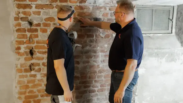 Zwei Männer untersuchen eine Ziegelwand. Ein Mann zeigt auf die Wand, die Anzeichen von Feuchtigkeit aufweist, ein Problem, das bei einem Haus ohne Bodenplatte auftreten kann.