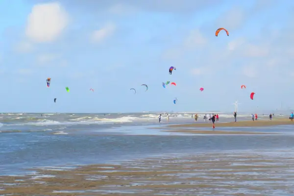 Plaża z wieloma latawcami do kitesurfingu na niebie i ludźmi spacerującymi po piasku.