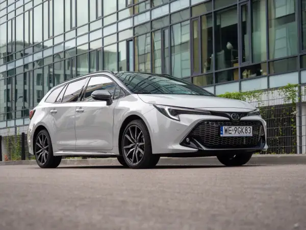 Biała Toyota Corolla 2.0 Hybrid kombi zaparkowana przed nowoczesnym budynkiem z lustrzaną fasadą.