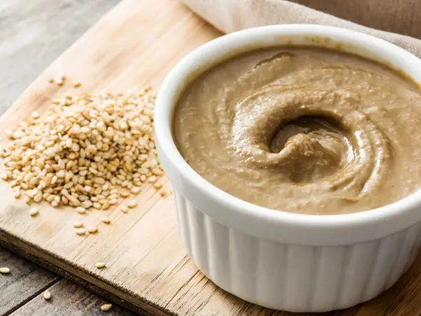 Un bol de sauce tahini crémeuse, à côté d'une pile de graines de sésame sur une planche en bois.