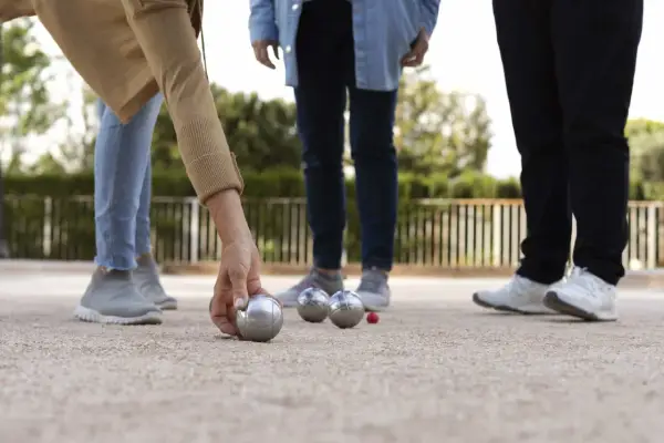 Une main de nom joueur de pétanque français saisit une boule argentée sur un terrain de gravier. D'autres boules et un cochonnet sont visibles.
