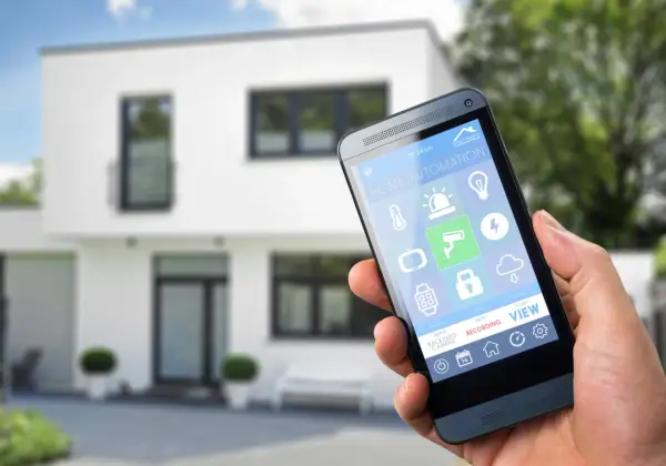 Dłoń trzyma smartfon z aplikacją do **instalacji inteligentnego domu**. Na ekranie widoczne ikony sterowania oświetleniem, ogrzewaniem, kamerami i innymi funkcjami.