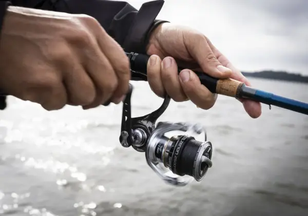 Händer som håller i ett fiskespö med en rulle, redo för fiska i Sverige. Vattnet glittrar i bakgrunden.