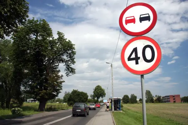 Znaki drogowe: zakaz wyprzedzania i ograniczenie prędkości do 40 km/h. Samochody jadą drogą, po prawej stronie przystanek autobusowy.