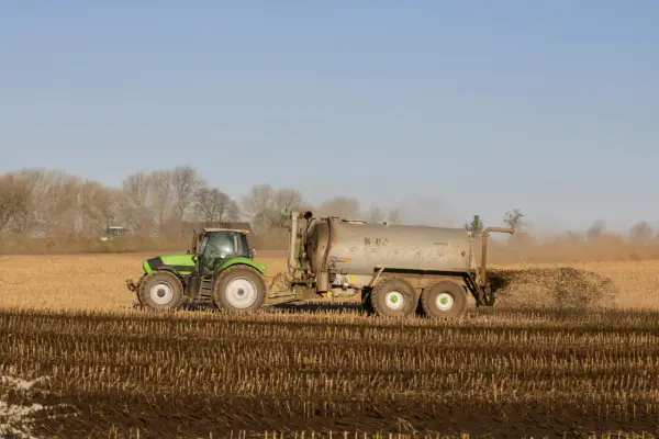 Zielony traktor z przyczepą rozprowadza obornik bydlęcy na polu.