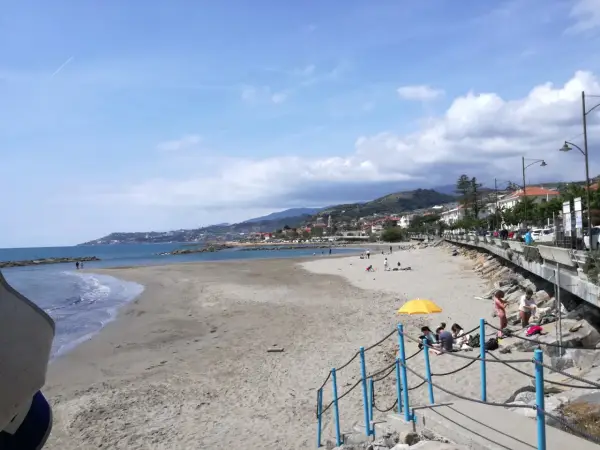Plage de sable à Santo Stefano al Mare, avec des gens profitant du soleil et de la mer. La ville s'étend le long de la côte, bordée de collines verdoyantes.