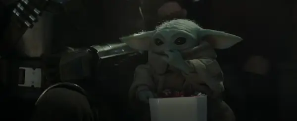 Dziecko z mandalorian film, w brązowym płaszczu, je słodycze z białego pudełka. Obok widać fragment zbroi Mandalorianina.