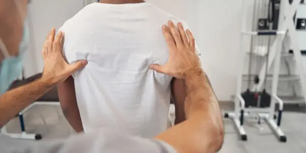 Fizjoterapeuta bada plecy pacjenta w białej koszulce.