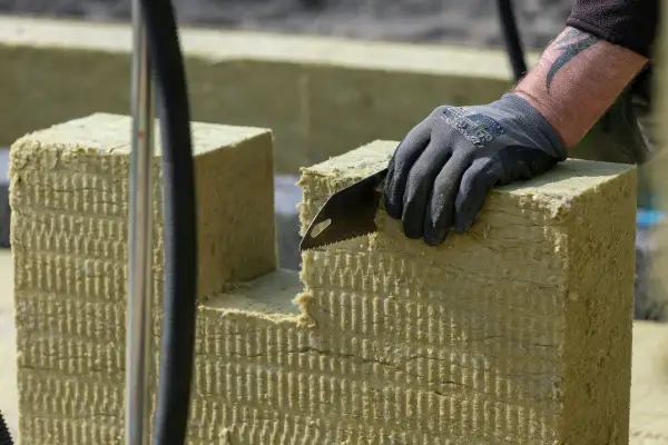 Ręka w rękawicy tnie piłą wełnę skalną 10. Materiał izolacyjny jest cięty na budowie.