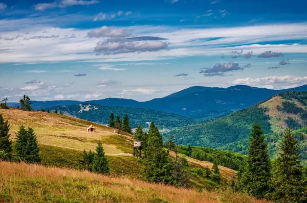 Widok na malowniczą **przełęcz Przegibek** z drewnianą chatką i wieżą widokową na zboczu. W tle gęsto zalesione góry pod błękitnym, chmurnym niebem.