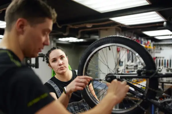 Mechanik rowerowy wyjaśnia, ile kosztuje wymiana dętki w rowerze, podczas gdy kobieta z zielonymi włosami uważnie słucha.