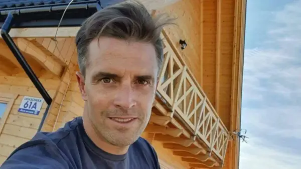 Staszek Karpiel-Bułecka pozuje na tle drewnianego domu z balustradą.