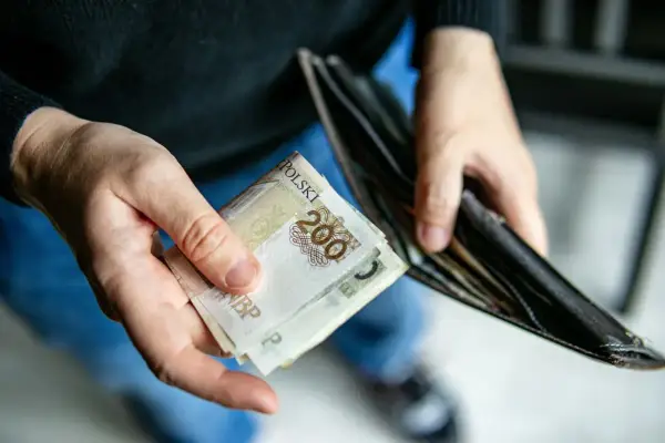 Dłonie trzymające polskie banknoty 200 zł i pusty portfel. Może to symbolizować trudności finansowe lub oczekiwanie na świadczenie pielęgnacyjne.