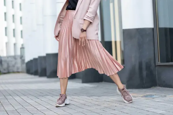 Kobieca stylizacja: pudrowy różowy żakiet i plisowana spódnica. Jakie buty do plisowanej spódnicy? Sneakersy w tym samym kolorze tworzą modny look.