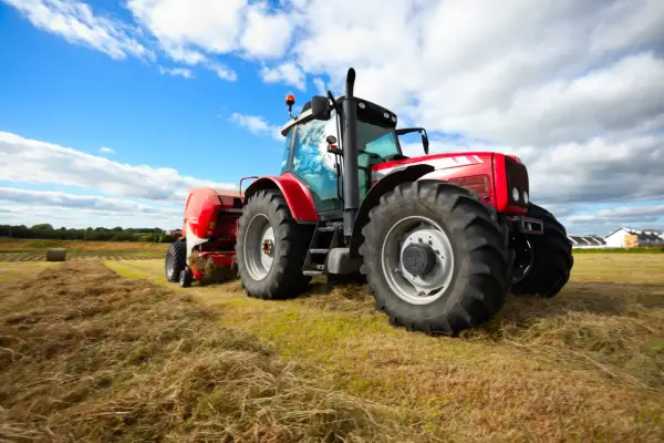 Czerwony traktor z przyczepą prasuje siano na polu. W tle błękitne niebo z chmurami. Traktorzysta w pracy.