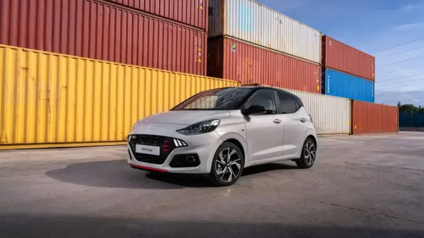Szary Hyundai i10 N Line z czerwonymi akcentami na grillu i zderzaku, zaparkowany przed stosem kolorowych kontenerów. Sprawdź atrakcyjną hyundai i10 cena!