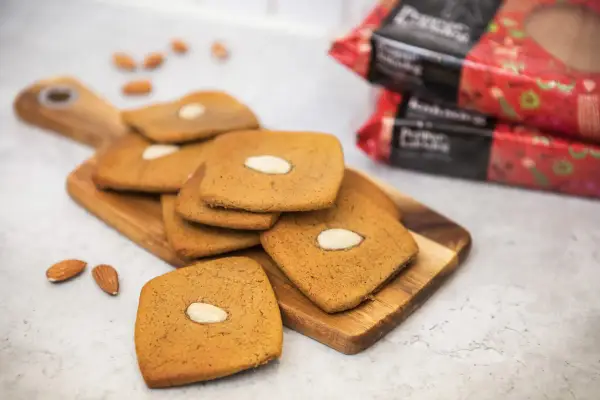 Fler fyrkantiga pepparkakor med mandel ligger på en träskärbräda, med några lösa mandlar och förpackningar i bakgrunden.
