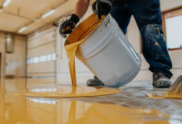 Hombre con guantes vierte pintura para suelo de cemento de color amarillo brillante de un cubo.